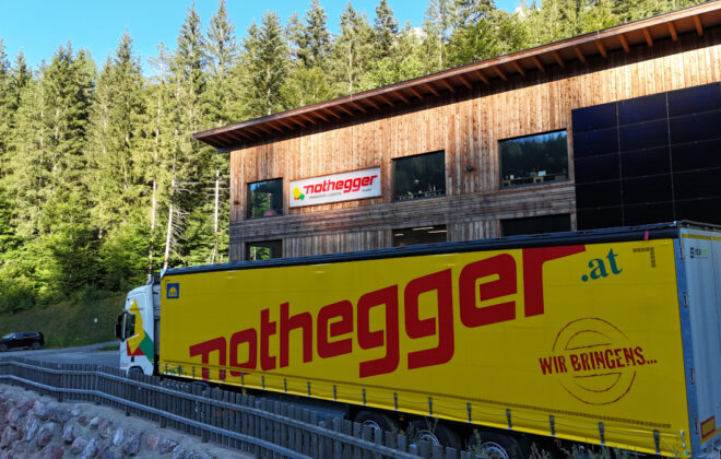 Sattelzug mit gelber Plane und Nothegger Schriftzug steht vor der Firmenzentrale, einem modernen Holzgebäude ebenfalls mit Nothegger Schriftzug in St. Ulrich am Pillersee